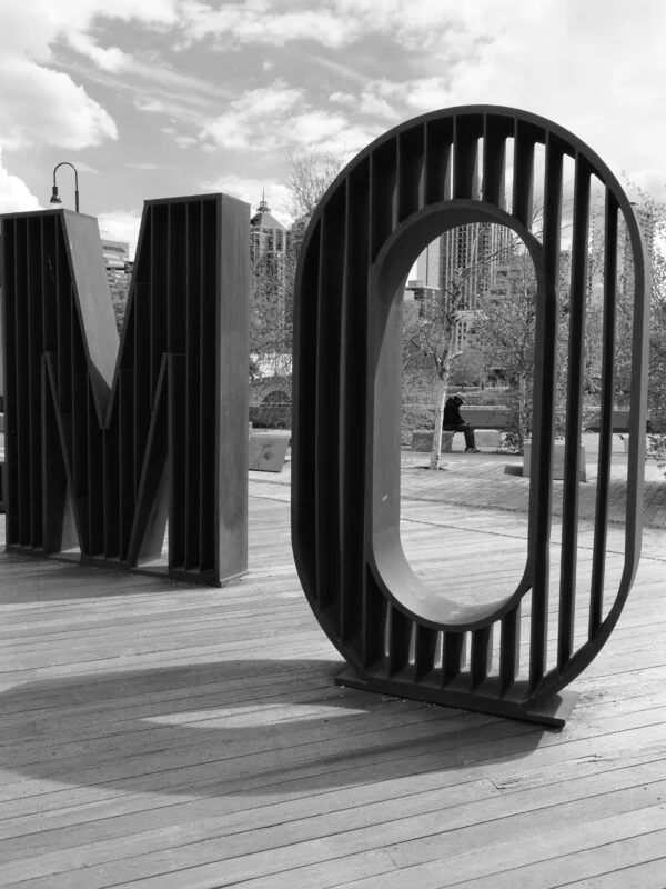 Великі металеві літери "M" та "O" стоять вертикально на дерев'яній палубі у відкритому міському парку, з деревами та високими будівлями на задньому плані. Зображення в чорно-білому кольорі.