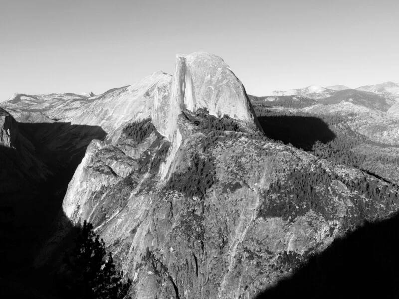Чорно-білий пейзажний вид на круту гранітну стіну Half Dome, що здіймається над лісистим гірським хребтом.