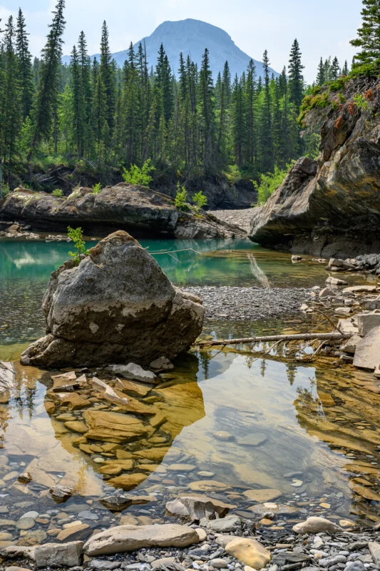 Спокійний гірський пейзаж з чистими, мілкими водами на передньому плані, що відкривають гладкі камені під ними. Великий камінь стоїть у воді, а високі сосни оточують берег. На задньому плані височіє величний гірський пік під частково хмарним небом.